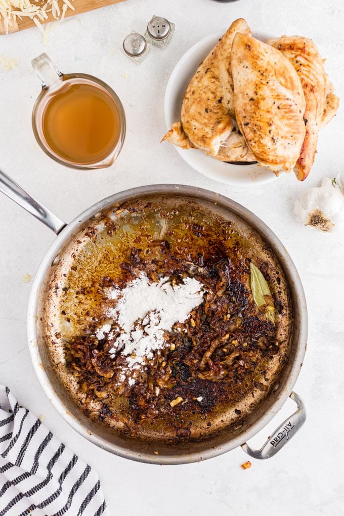 Skillet with caramelized onions and flour, plate of seared chicken breasts, beef broth, striped linen, garlic clove, on a white marble countertop.