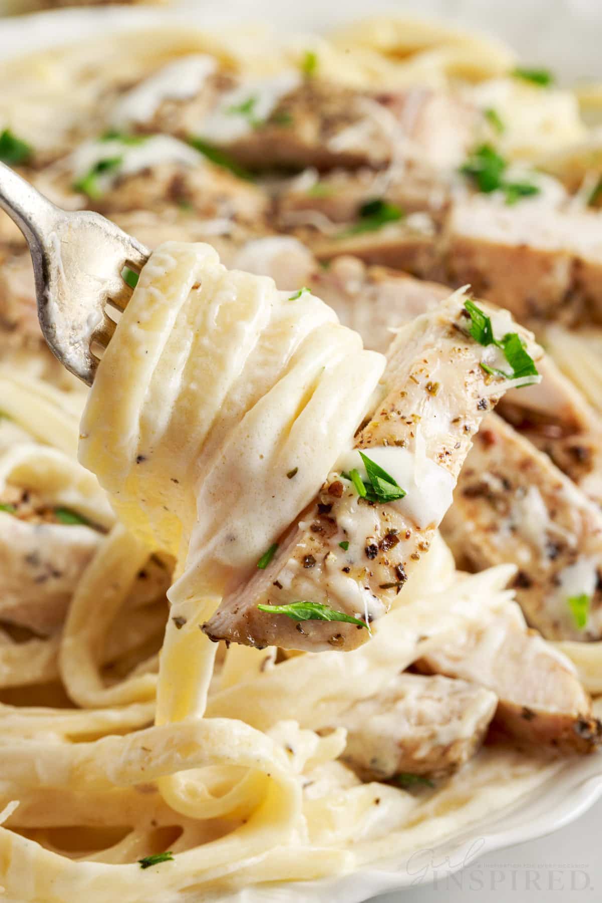 a spoon with fettuccine wrapped around it with a piece of chicken on the end over a plate of grilled chicken alfredo