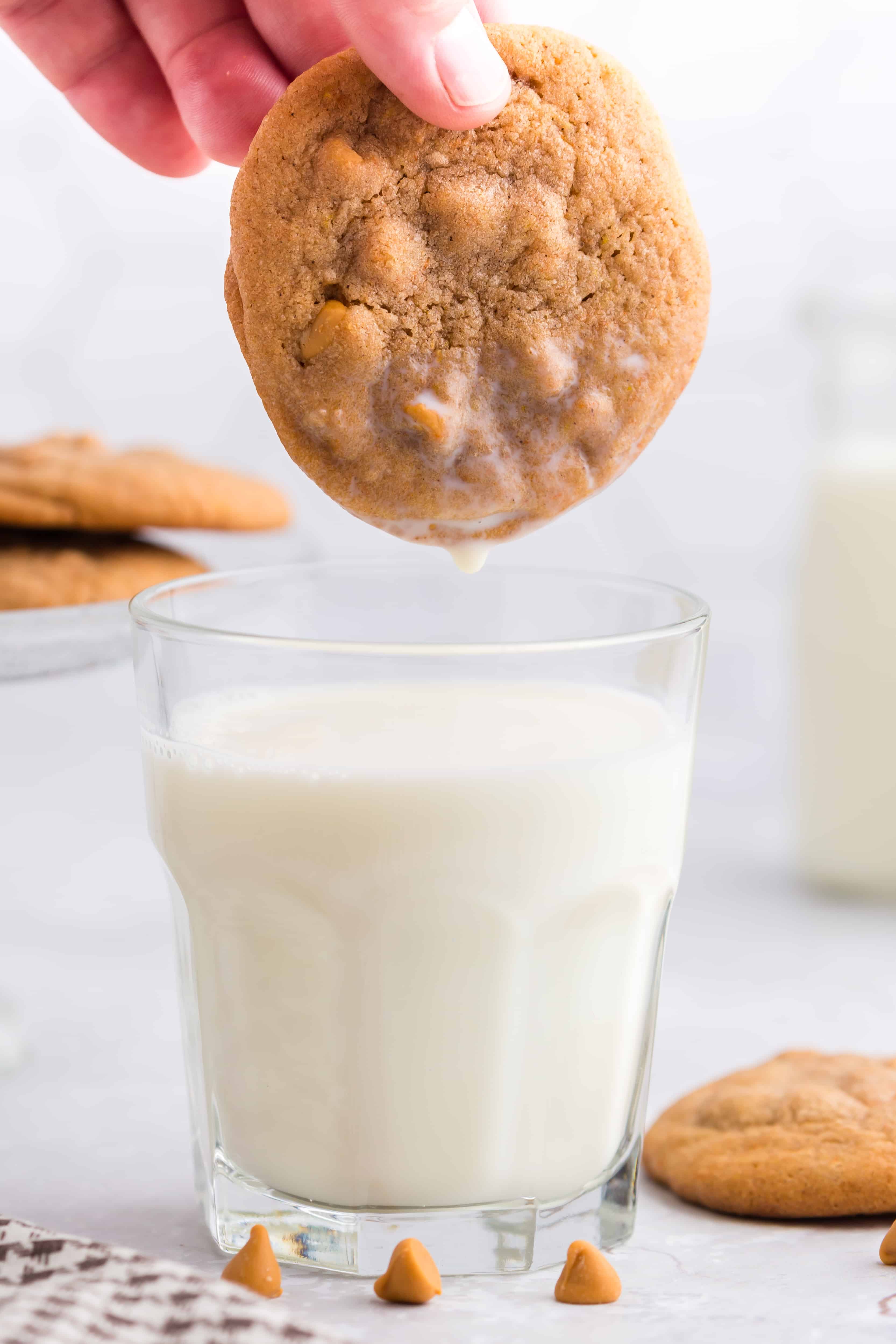 Hand dipping a pumpkin butterscotch cookie into a glass of milk, glass cake stand with cookies, checkered linen, jug of milk, single pumpkin butterscotch cookie with loose butterscotch chips on a grey marble countertop.
