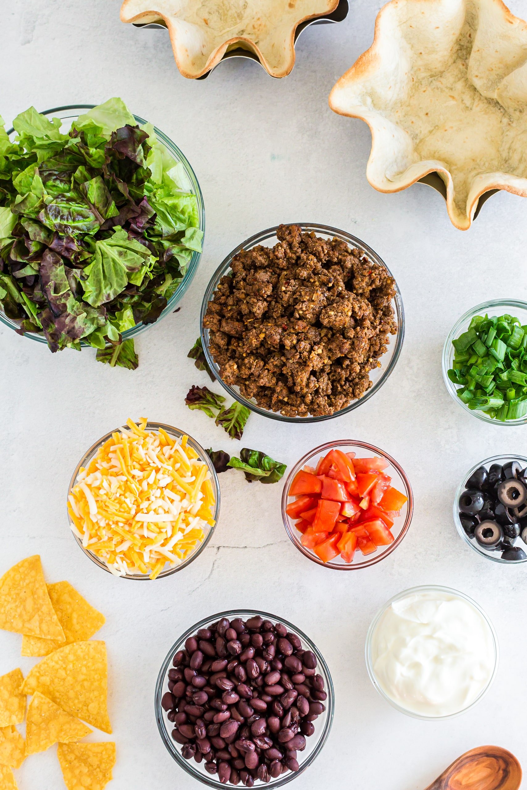 individual ingredients for taco salad bowls set out like lettuce, taco meat, cheese, diced tomatoes, etc.