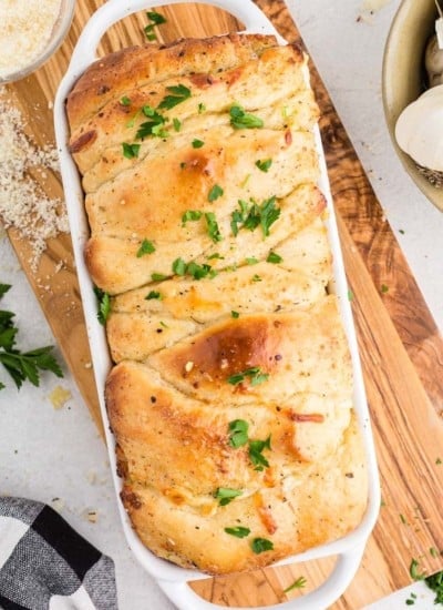 Close up of freshly baked pull apart garlic bread in white bread loaf baking dish on wooden kitchen board, black and white checkered linen, bowl of whole garlic cloves, on a white marble surface.