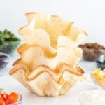 A stack of four flour tortillas baked into fluted taco salad shells, with taco meat, tomatoes, lettuce, and cheese in nearby bowls.