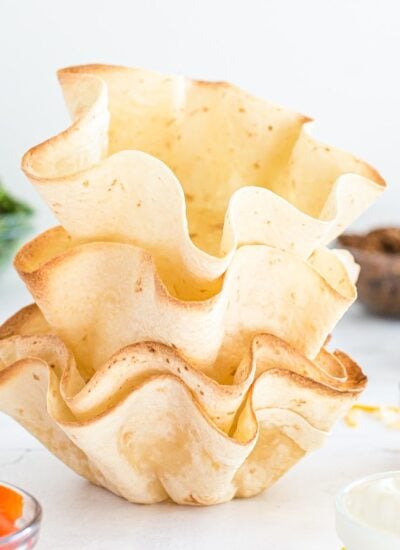 A stack of four flour tortillas baked into fluted taco salad shells, with taco meat, tomatoes, lettuce, and cheese in nearby bowls.