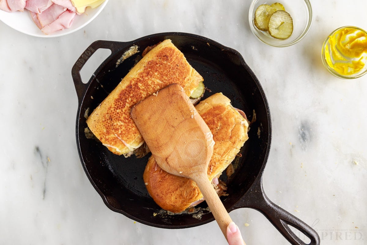 browned cheesecake factory cuban sandwiches in cast iron skillet with wooden spatula on top