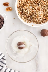 chocolate turtle thumbprint cookie dough ball in bowl of frothy egg whites next to crushed pecans