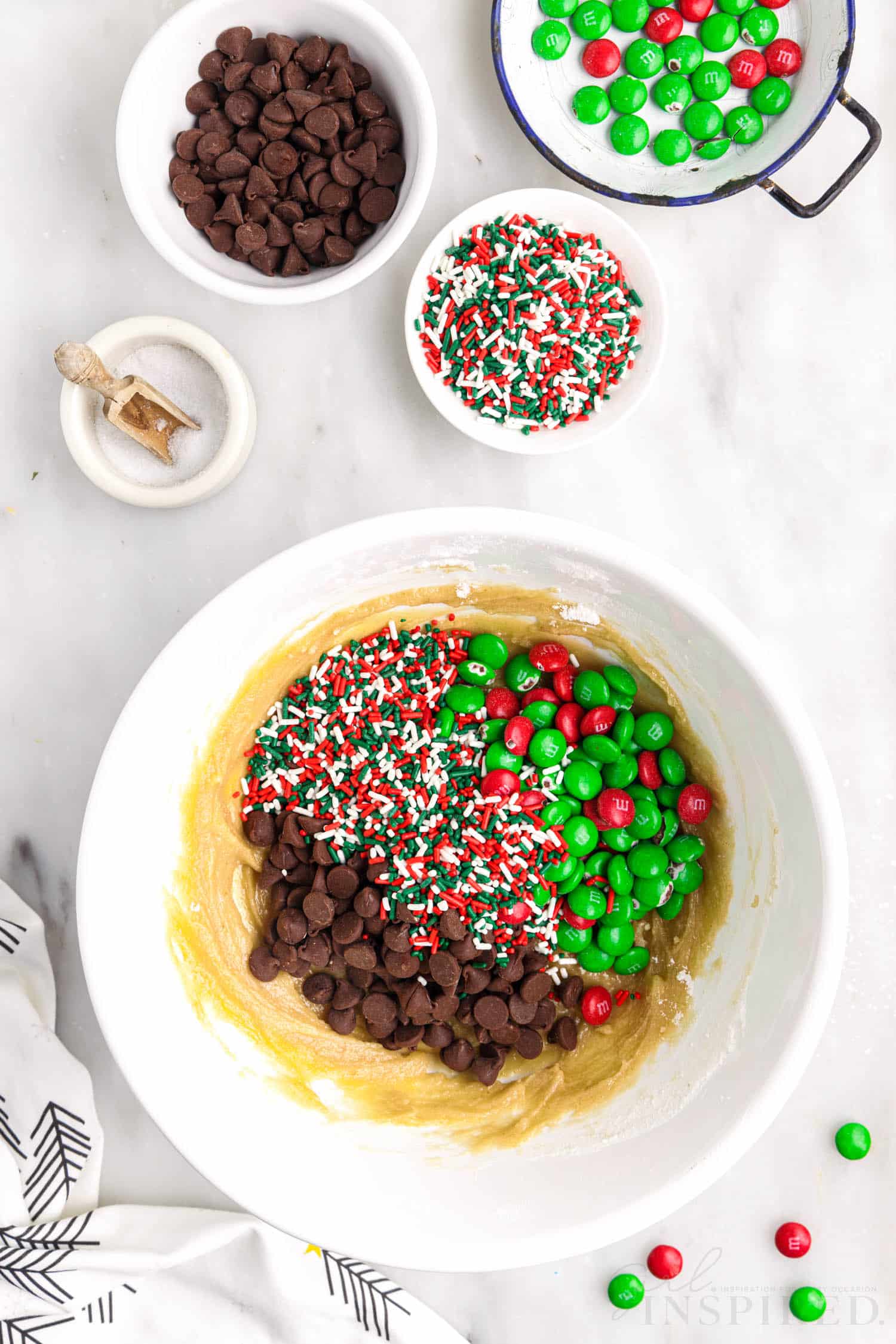 m&ms sprinkles and chocolate chips added to egg flour mixture in a white mixing bowl