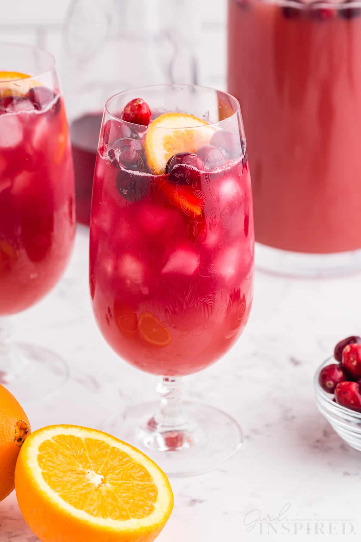 Two glasses of Christmas punch made using the Christmas punch recipe (non alcoholic), on a marble countertop, halved fresh orange, pitcher with Christmas punch, bowl of cranberries, white tiled background.