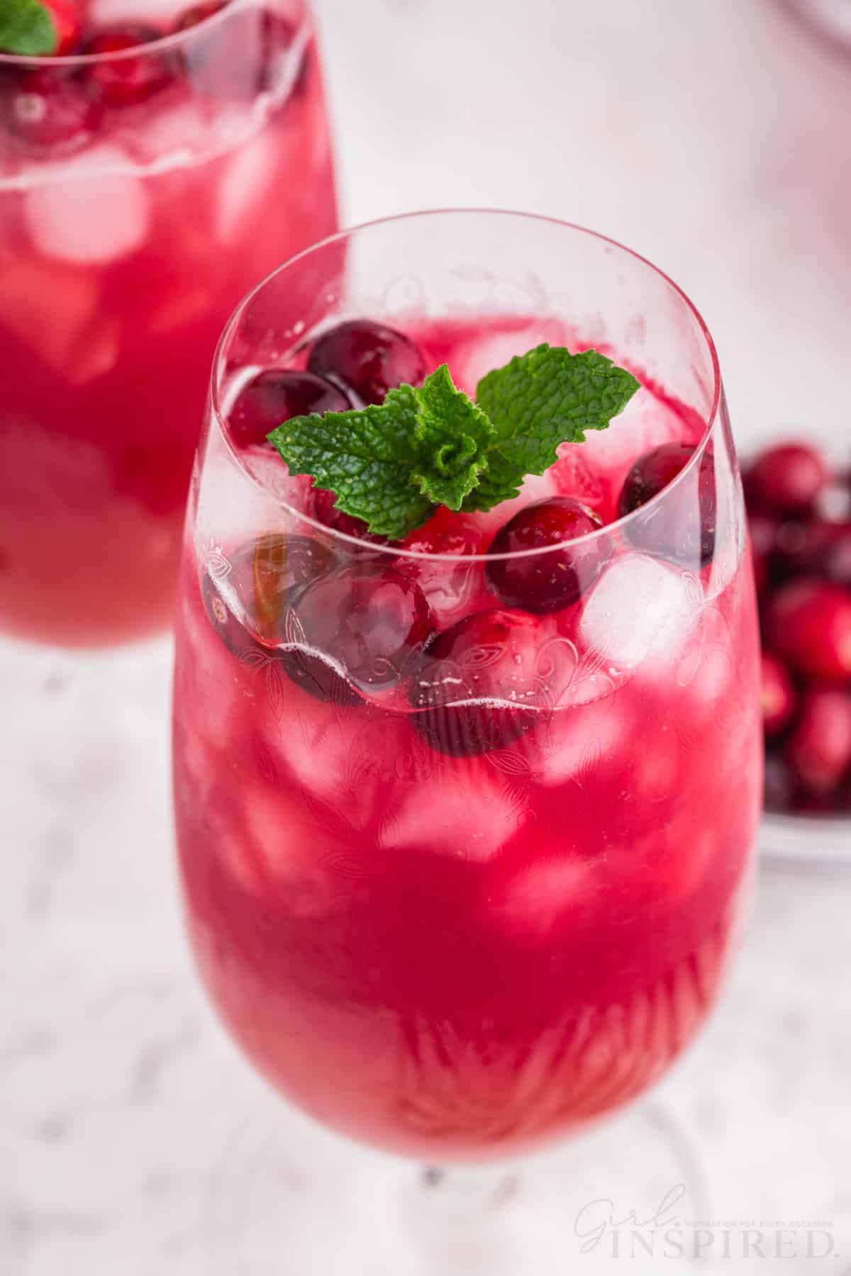 Two glasses with ice and Christmas punch, garnished with fresh cranberries and mint leaves, bowl of cranberries in the background, on a marble countertop.