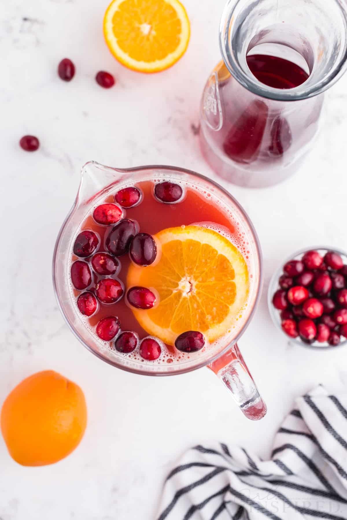 Overhead view of Christmas punch in a pitcher, striped linen, empty glass jug, bowl of cranberries, sliced orange, on a marble countertop.