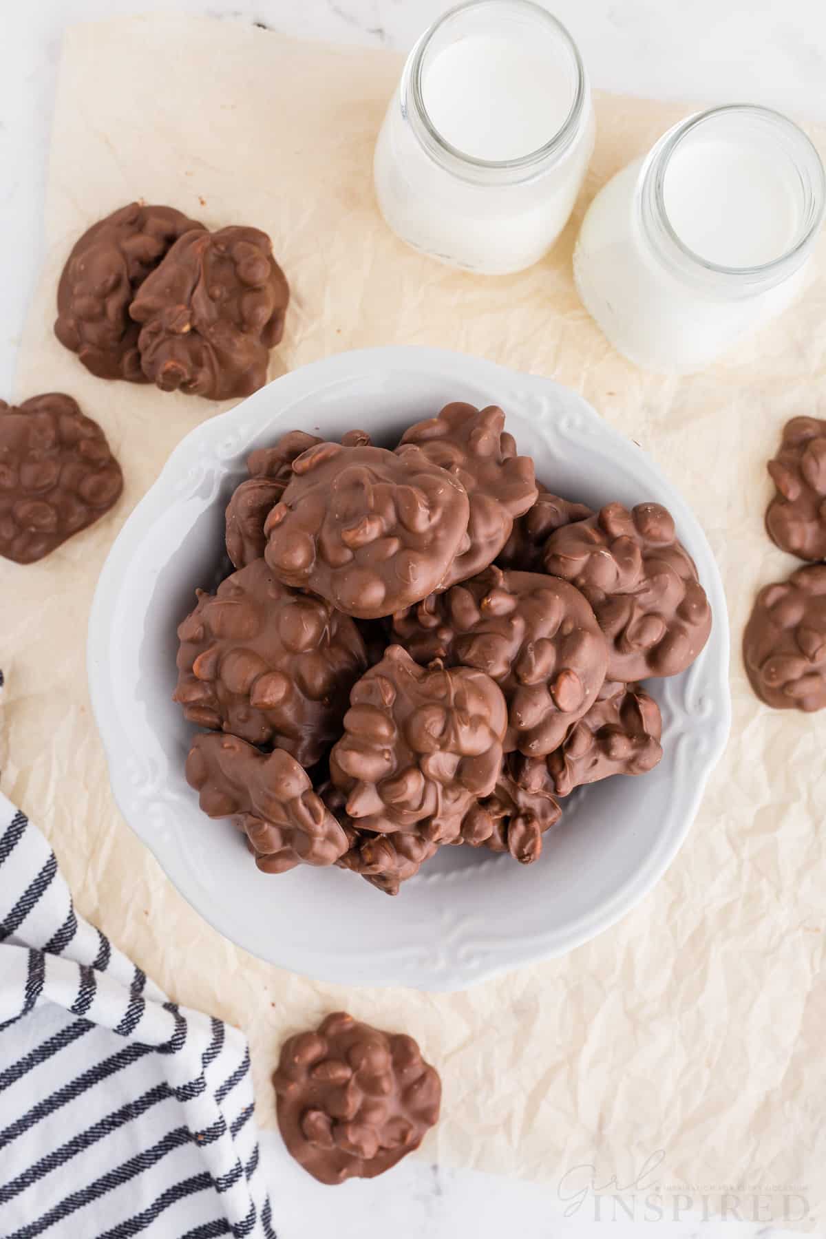 Overhead view of chocolate peanut clusters on a white cake stand, chocolate peanut clusters on a sheet of wax paper, two jars of milk, striped linen, on a marble countertop.