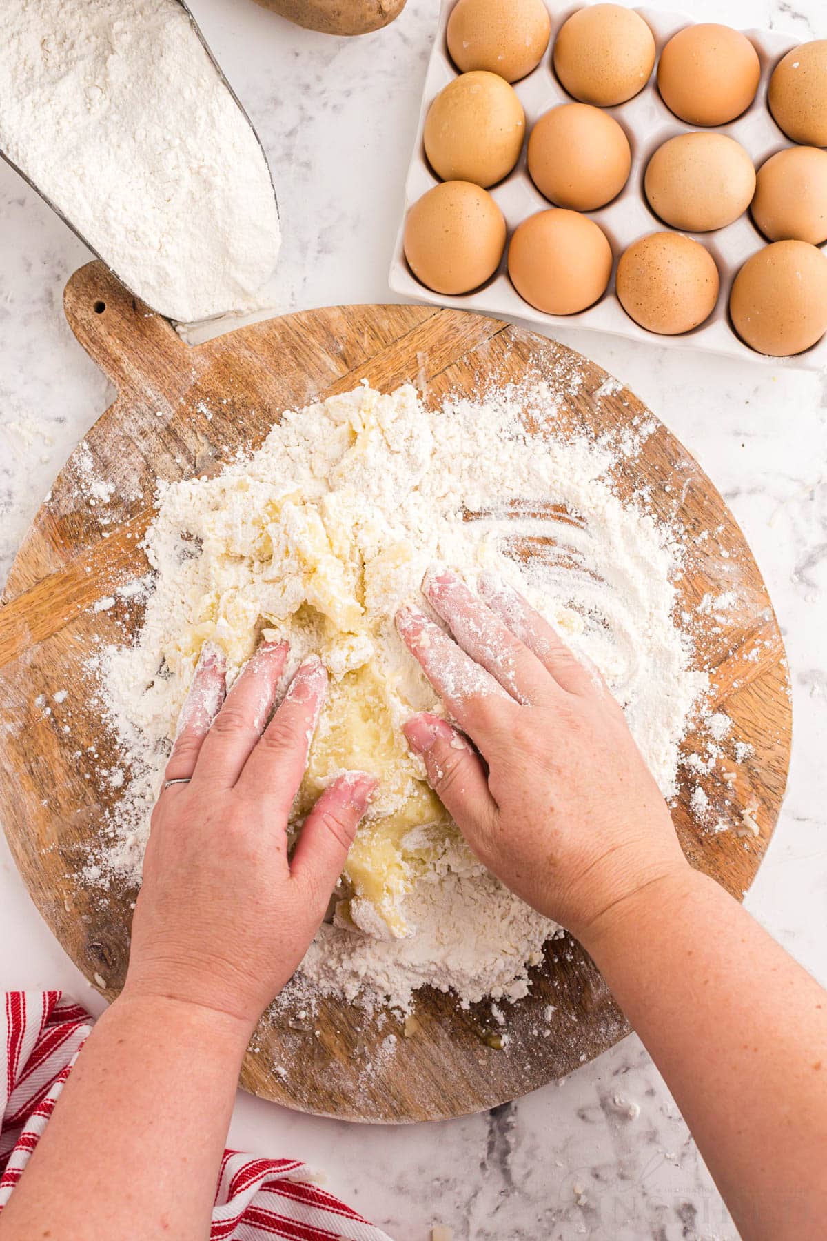 Hands mixing the mashed potato mixture with flour on a wooden kitchen board, tray of eggs, measuring spoon with flour, red and white striped linen, on a marble countertop.