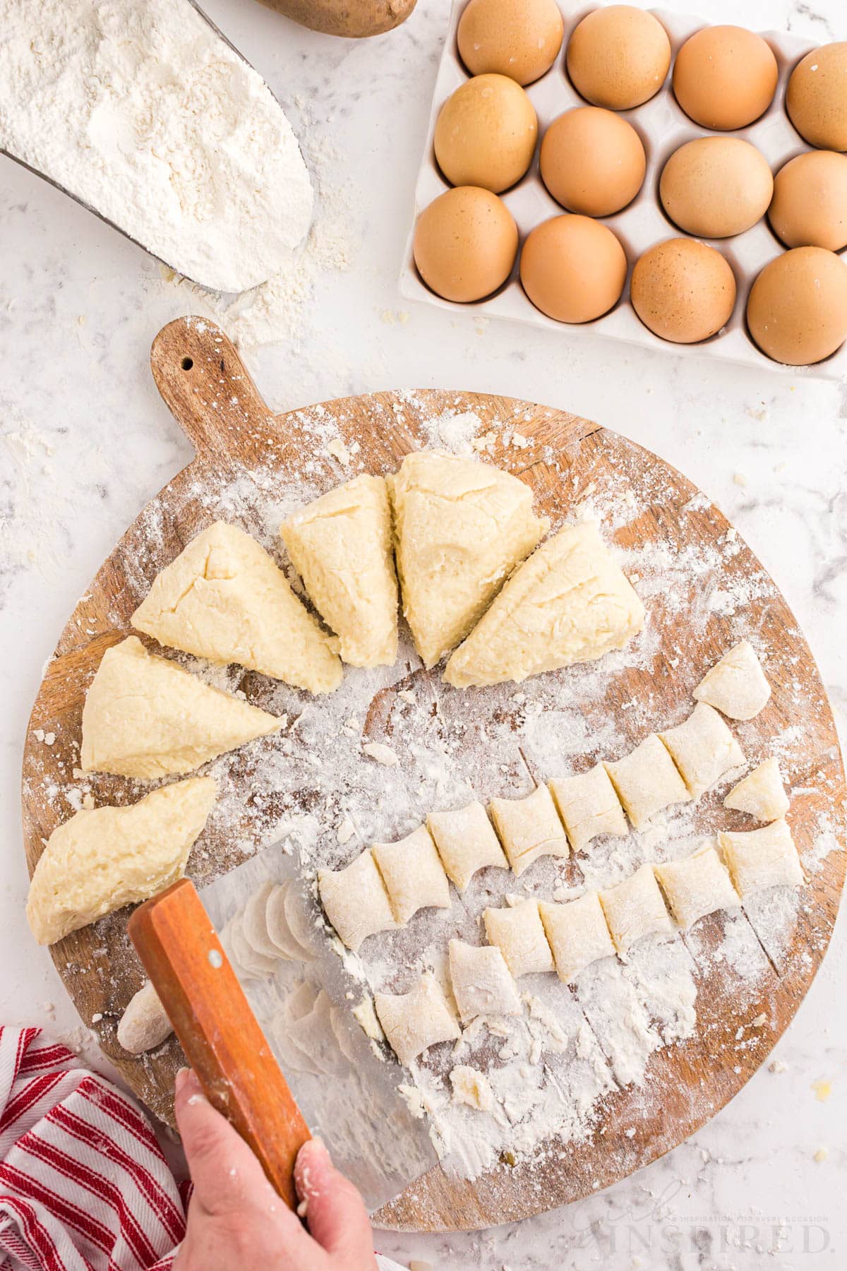 Wooden kitchen board with cut pieces of gnocchi dough, two rolls of gnocchi dough cut into equal pieces, red and white striped linen, tray of eggs, measuring spoon with flour, on a marble countertop.