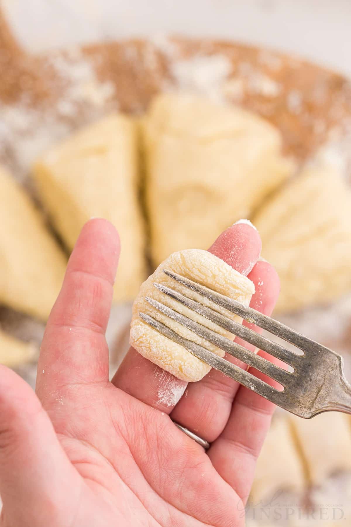 Metal fork pressed against piece of gnocchi dough against finger, gnocchi dough on a wooden kitchen board in the background.