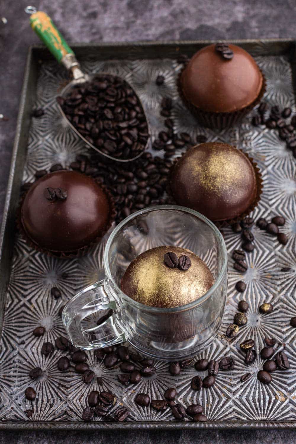 mocha hot chocolate bomb in a glass mug on a cookie sheet with a scoop of coffee beans next to additional mocha hot chocolate bombs