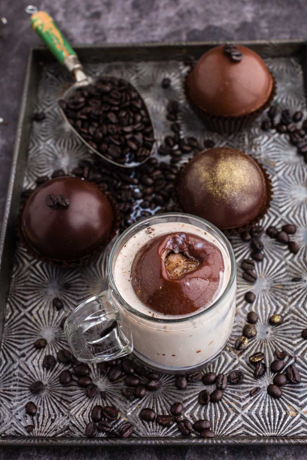 a cup of milk with a mocha hot chocolate bombs on a cookie sheet with coffee beans and three other mocha hot chocolate bombs