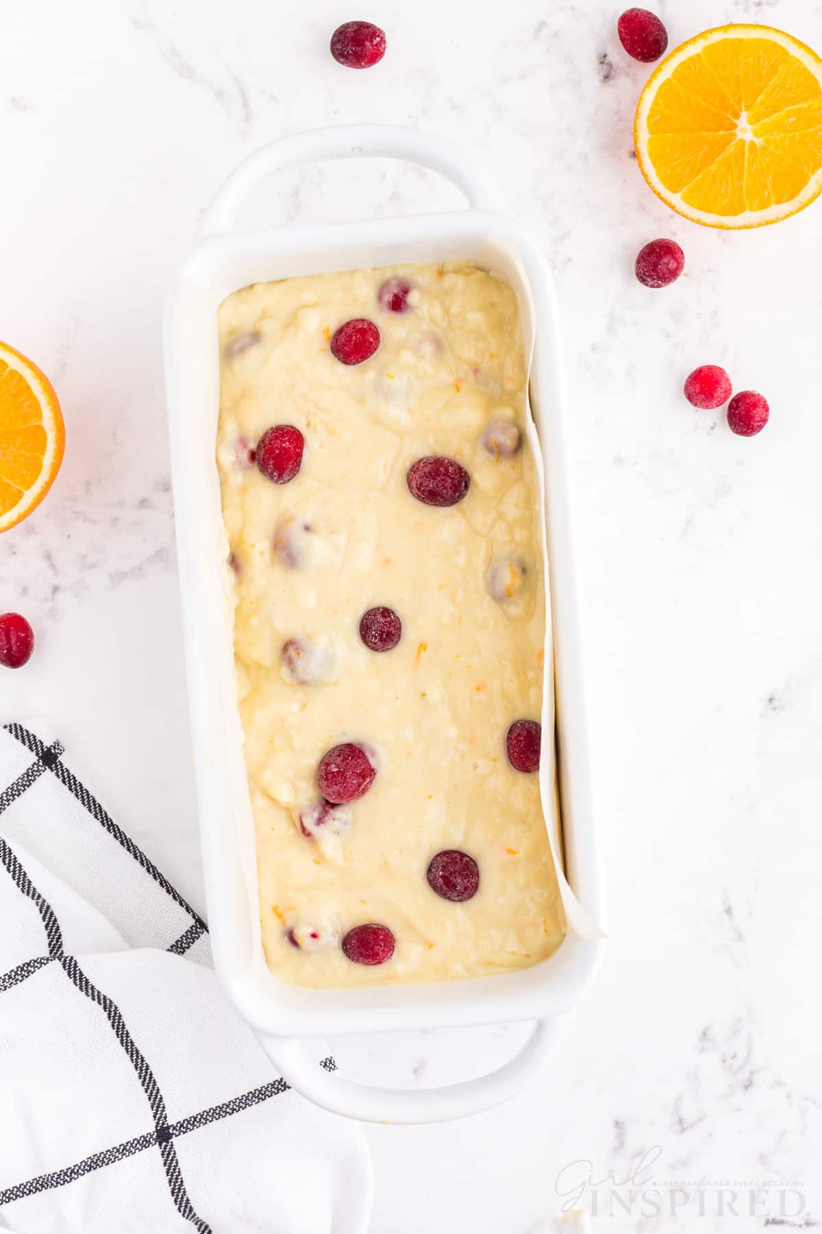 White prepared loaf pan with orange cranberry bread batter on a white marble countertop, checked linen, sliced oranges, whole loose cranberries.