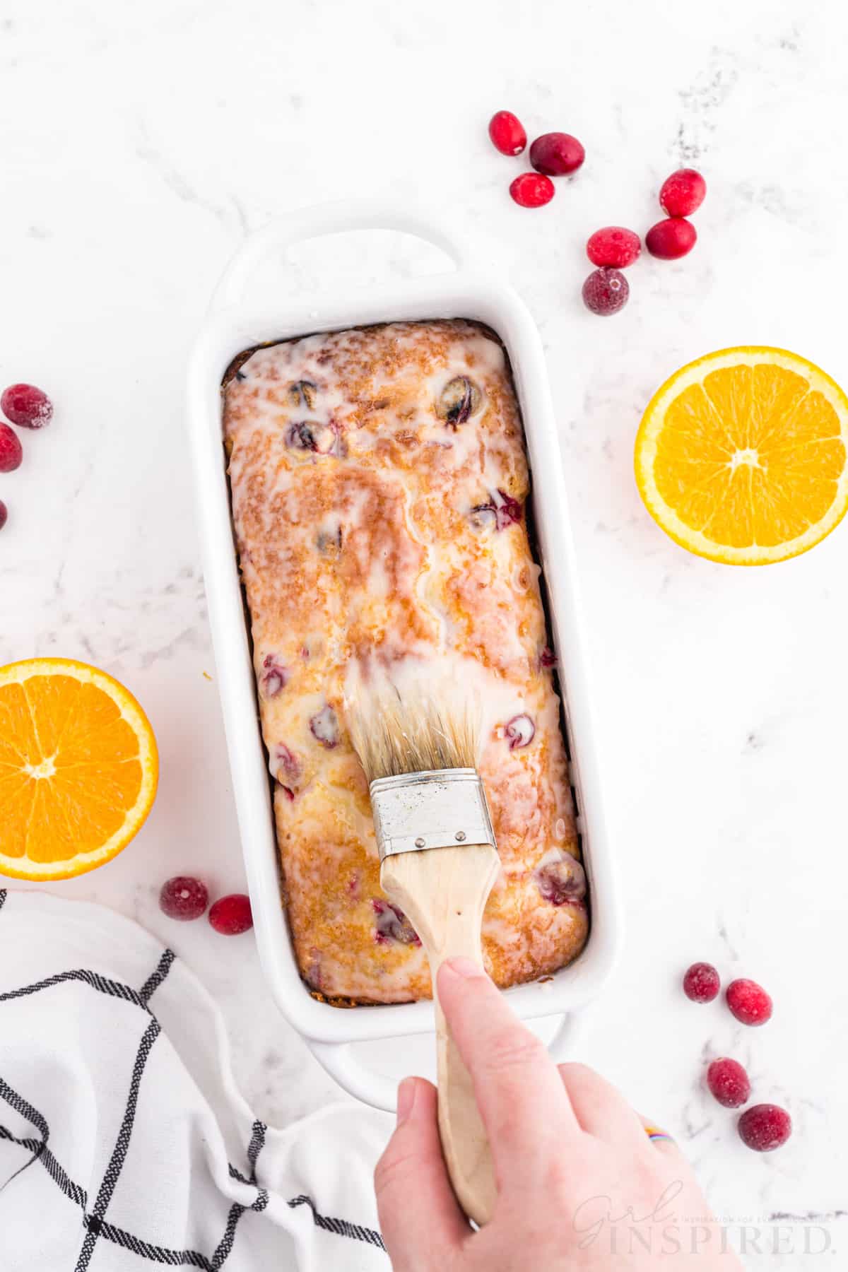 Hand brushing orange sugar glaze with a pastry brush on the top of the baked and cooled orange cranberry bread in white loaf pan, checked linen, sliced oranges, whole loose cranberries, on a white marble countertop.