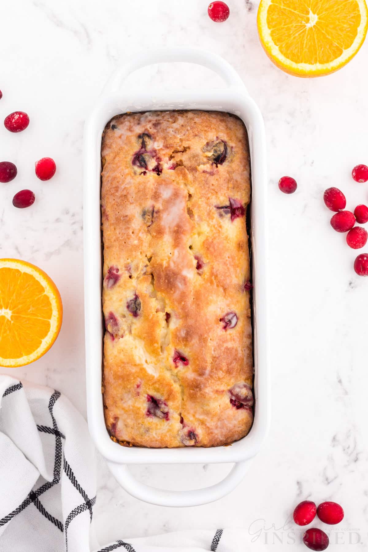 Baked Orange Cranberry Bread in white loaf pan, checked linen, sliced fresh oranges, fresh whole cranberries on a white marble countertop.