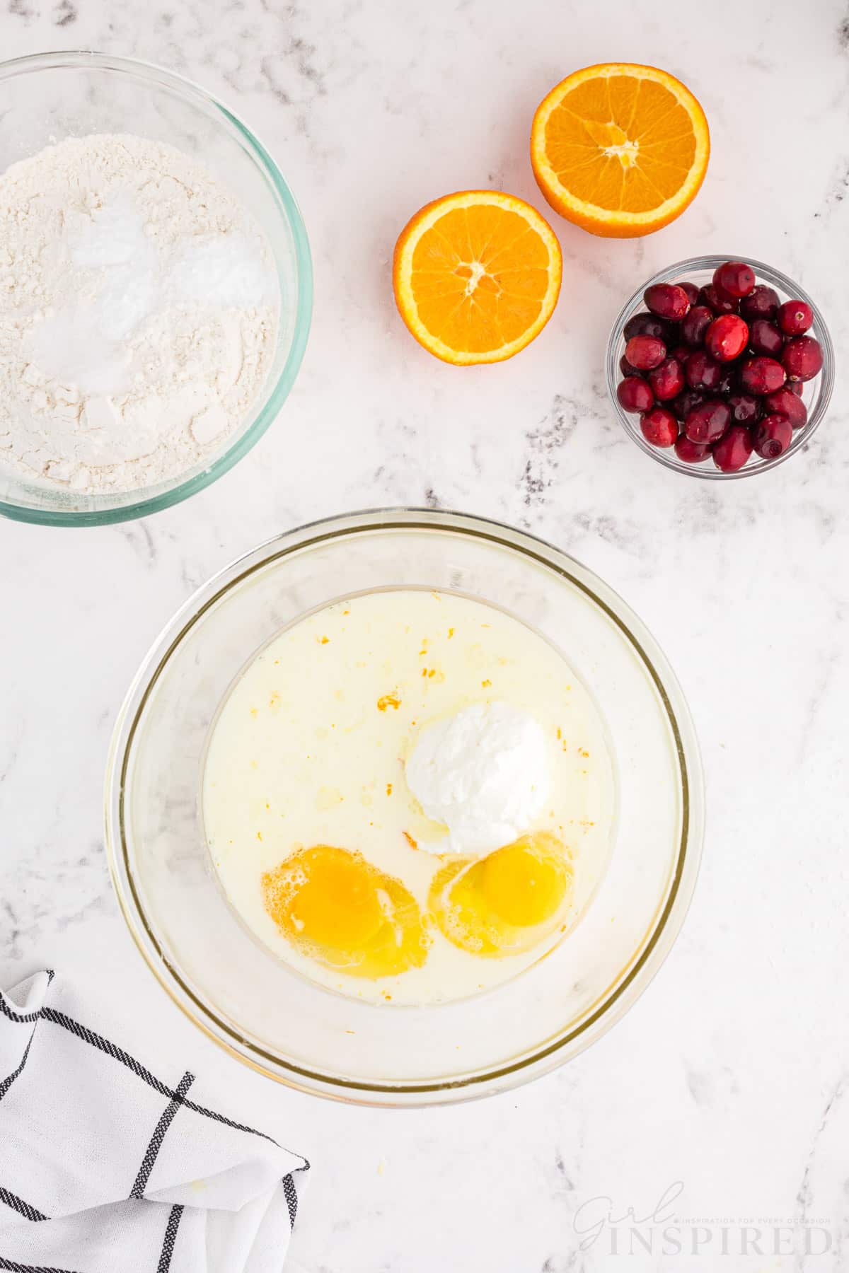 Mixing bowl with sugar, orange zest, eggs, buttermilk, and oil, bowl of whole cranberries, bowl of dry ingredients, fresh sliced oranges, checked linen on a white marble countertop.