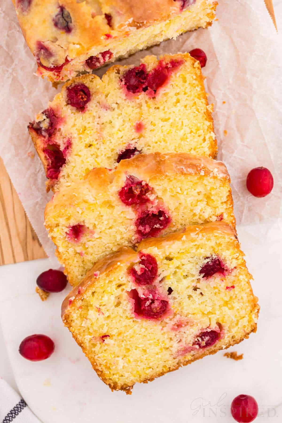Sliced orange cranberry bread on parchment paper on a wooden kitchen board, checked linen, fresh whole cranberries on a white marble countertop.