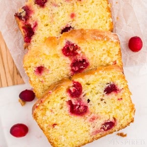 Baked and sliced orange cranberry bread on parchment paper on top of a wooden kitchen board on a white marble countertop with loose fresh cranberries.