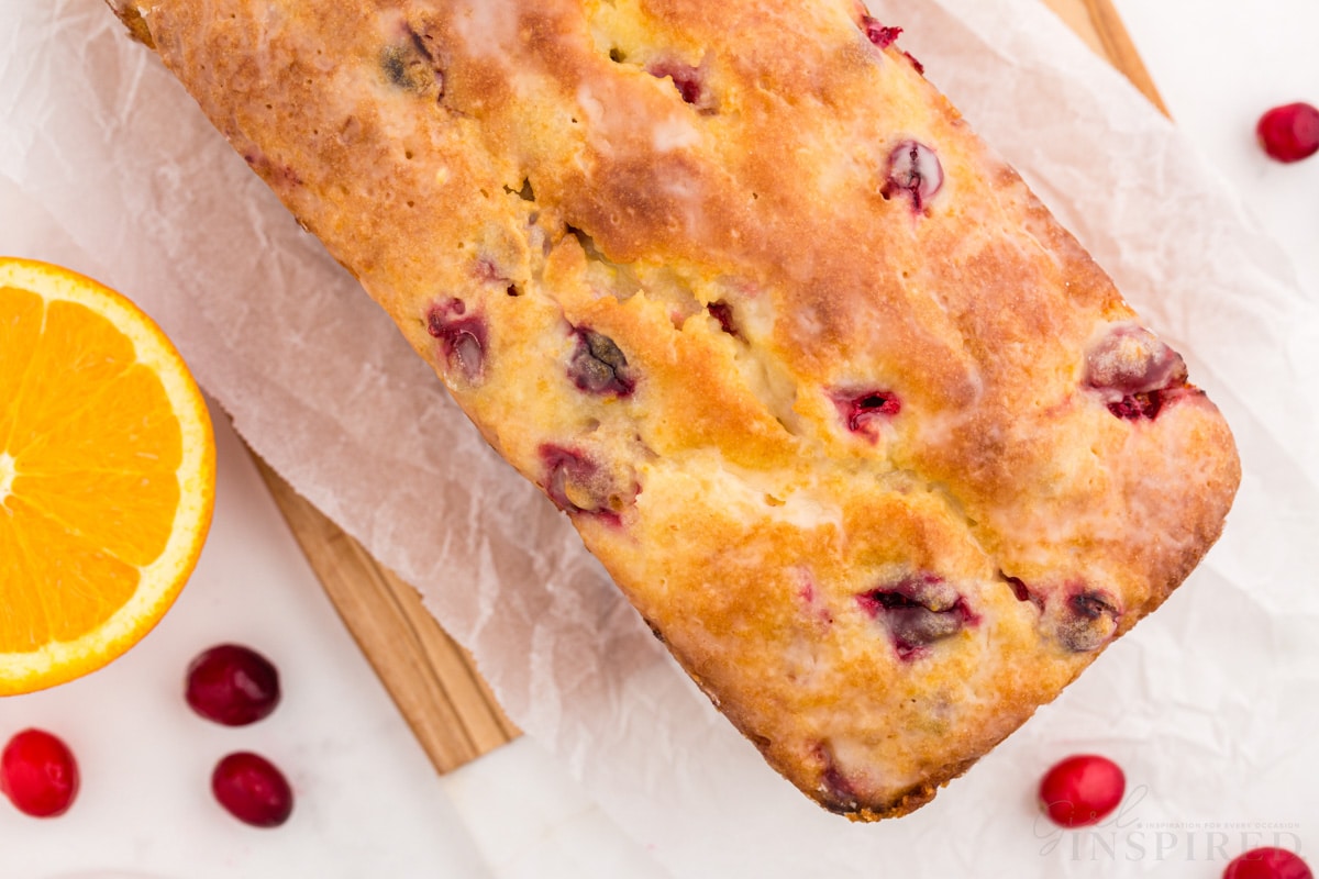 Baked orange cranberry bread on parchment paper on a wooden kitchen board on a white marble countertop, sliced orange and loose cranberries.