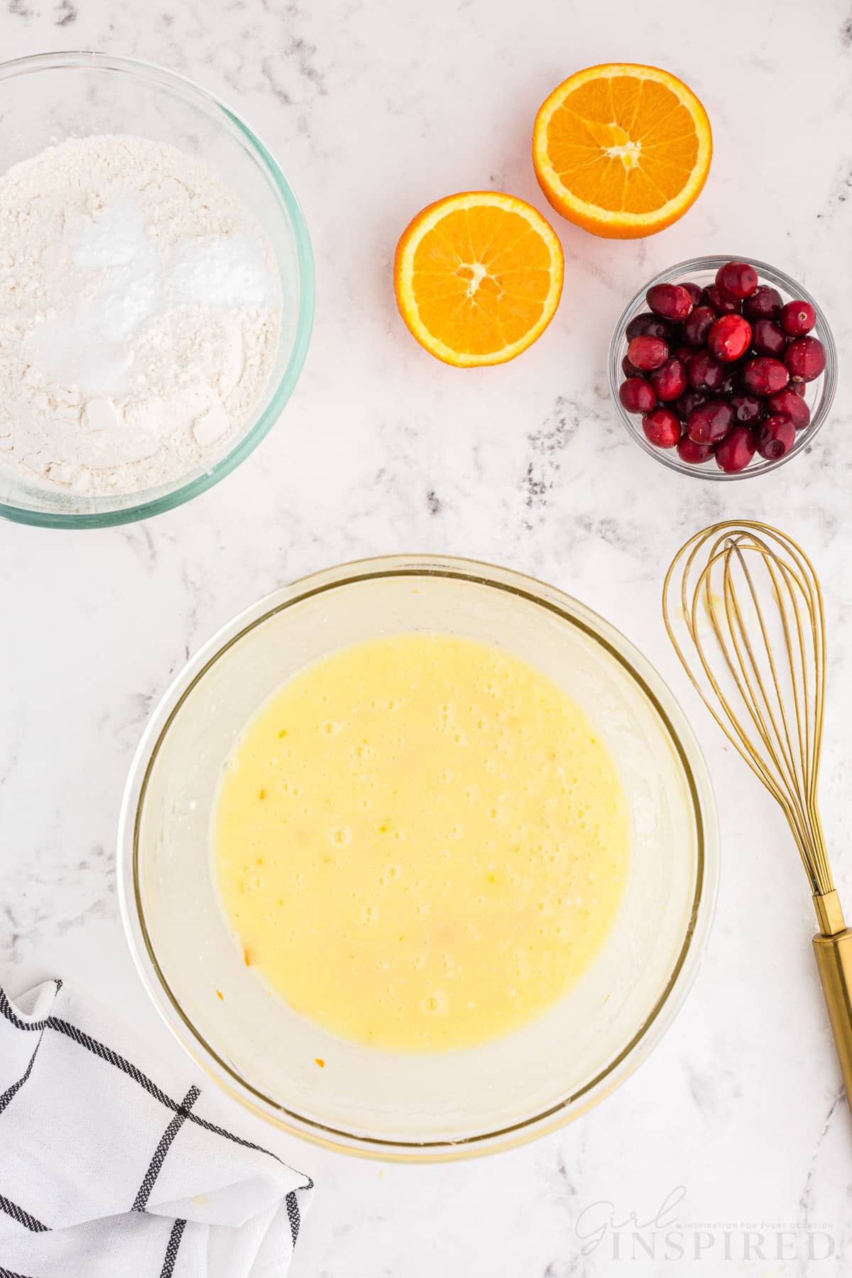 Mixing bowl with all wet ingredients combined with freshly squeezed orange juice, bowl of dry ingredients, bowl of fresh cranberries, sliced orange, checked linen, metal whisk on a white marble countertop.