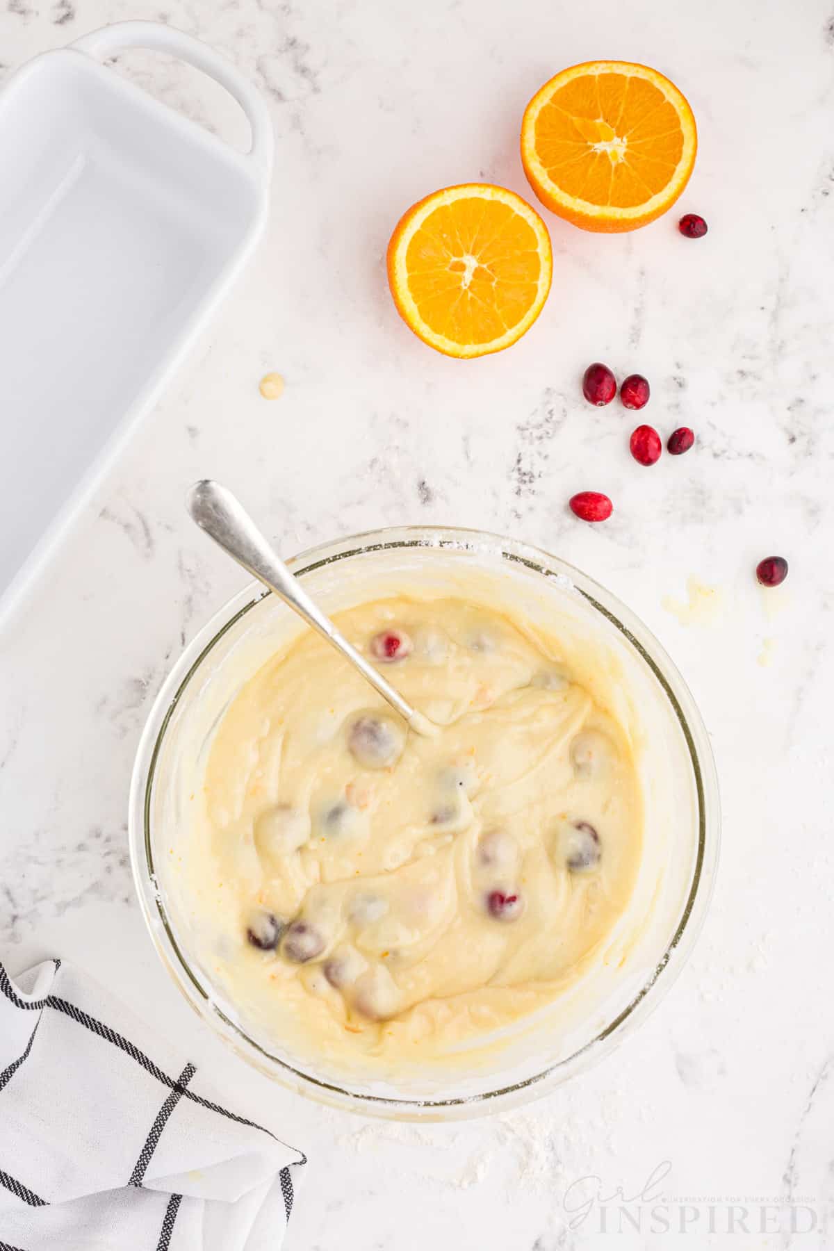 Bowl of orange cranberry bread with whole cranberries gently folded in the batter with metal spoon, white prepared loaf pan, checked linen, sliced orange, whole loose cranberries on a white marble countertop.