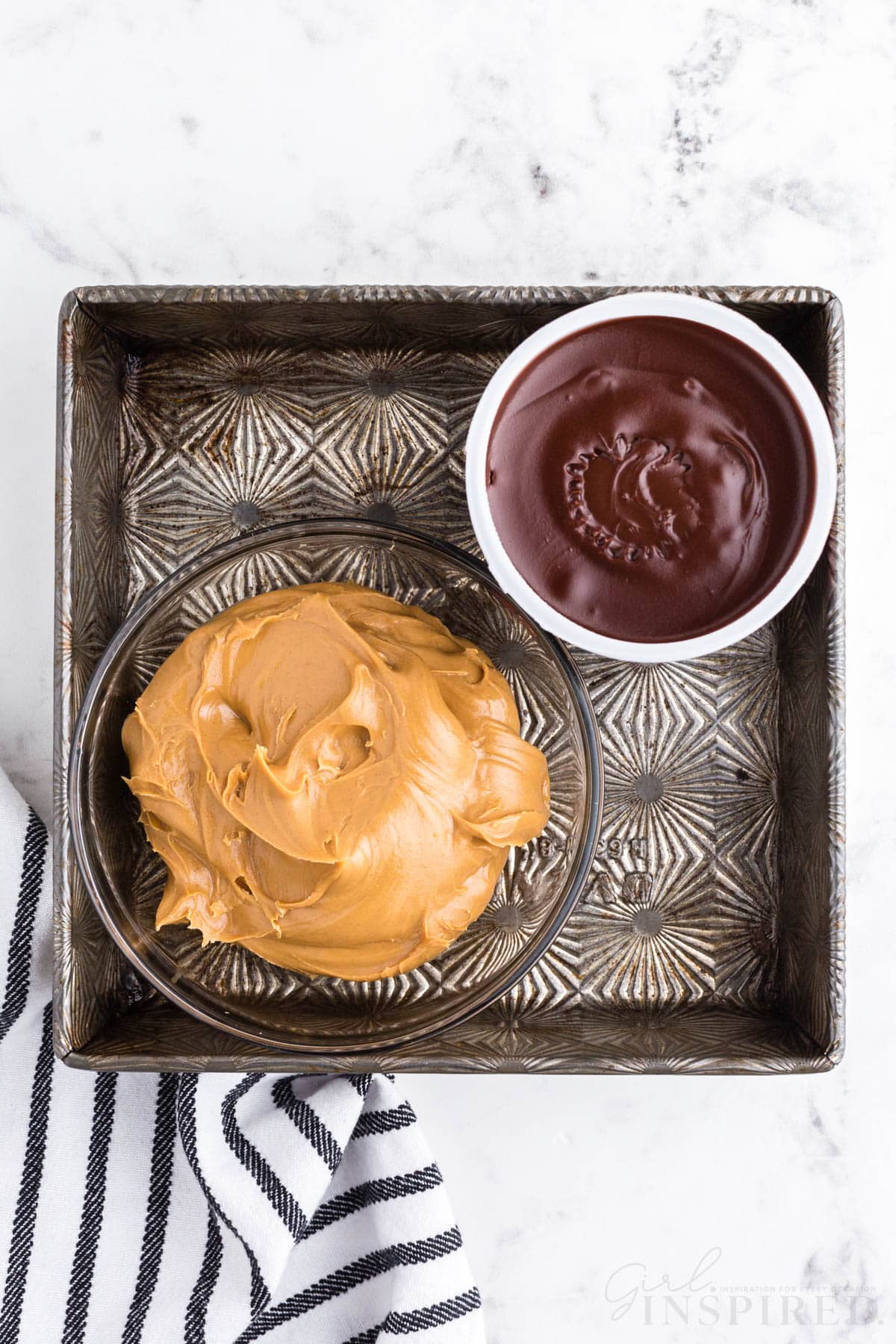 Metal baking tray with a bowl of creamy peanut butter and a bowl of chocolate frosting, striped linen, on a marble countertop.