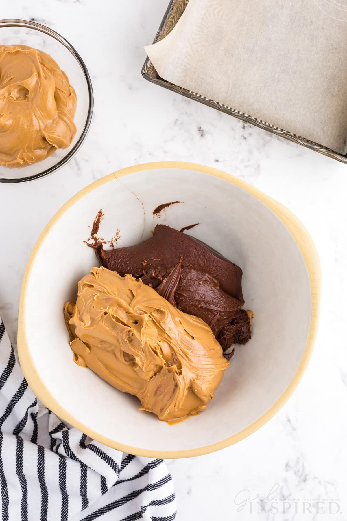 Mixing bowl with creamy peanut butter and chocolate frosting, glass bowl with remaining peanut butter, striped linen, metal baking tray lined with parchment paper, on a marble countertop.