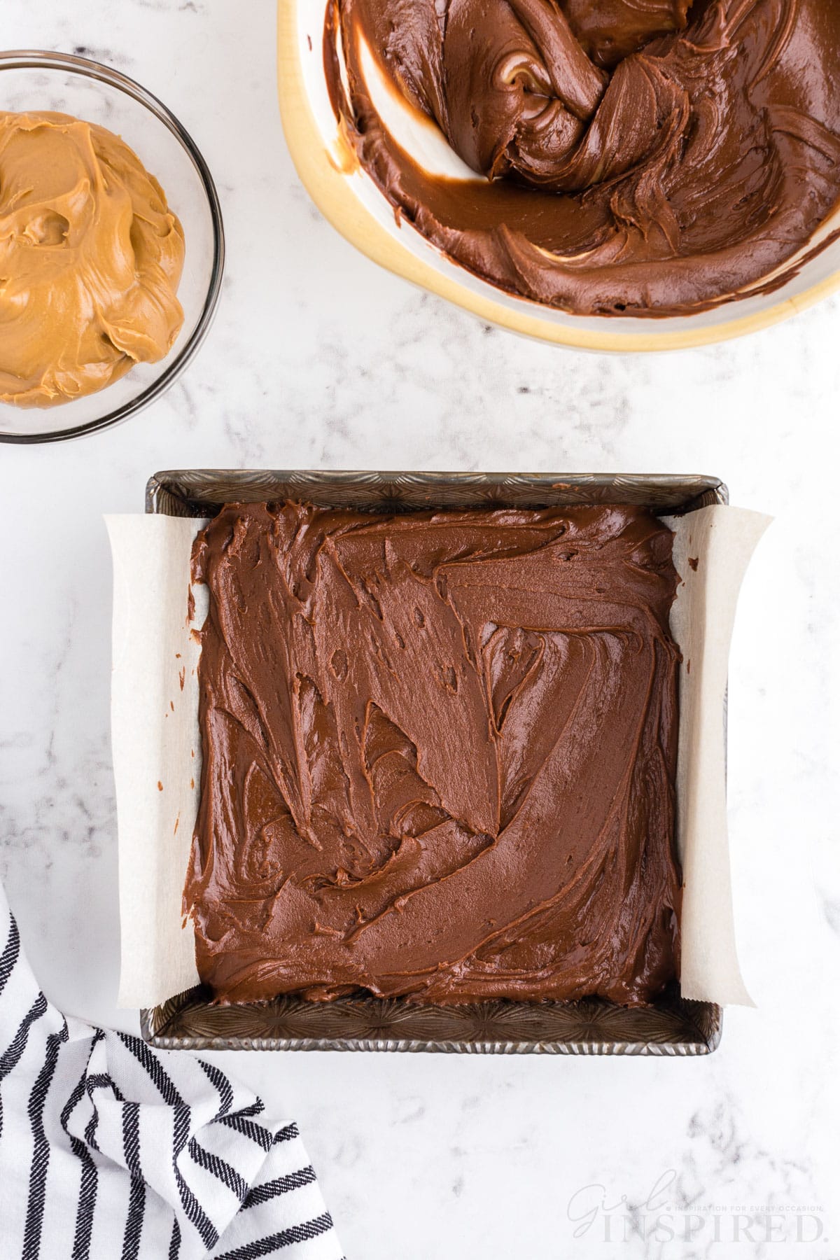 Baking tray lined with parchment paper and filled with some peanut butter and chocolate frosting mixture, glass bowl of remaining peanut butter, mixing bowl with remaining chocolate frosting and peanut butter mixture, striped linen, on a marble countertop.