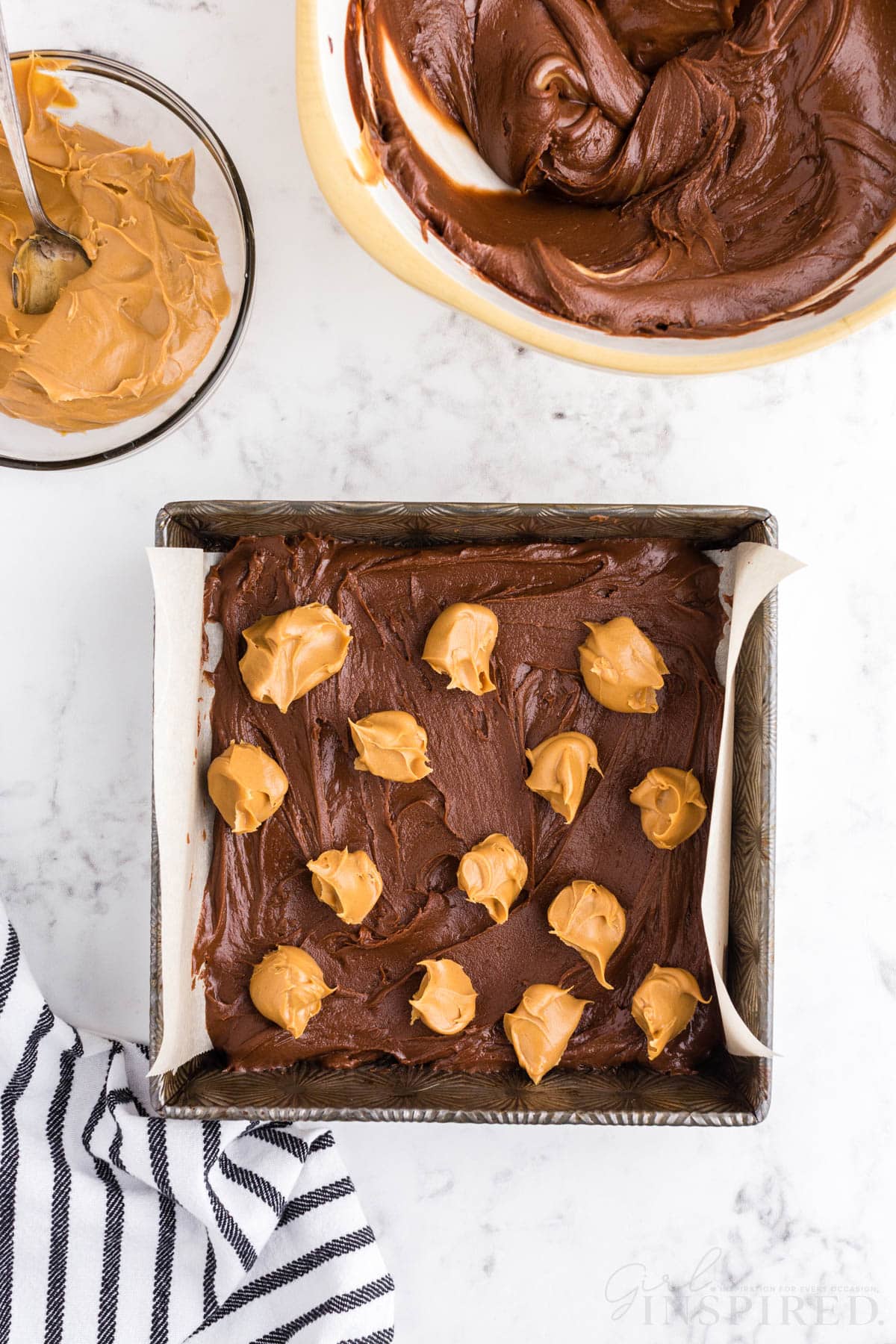 Baking tray with chocolate peanut butter fudge mixture and dollops of creamy peanut butter on top, glass bowl of creamy peanut butter, mixing bowl with fudge mixture, striped linen, on a marble countertop.