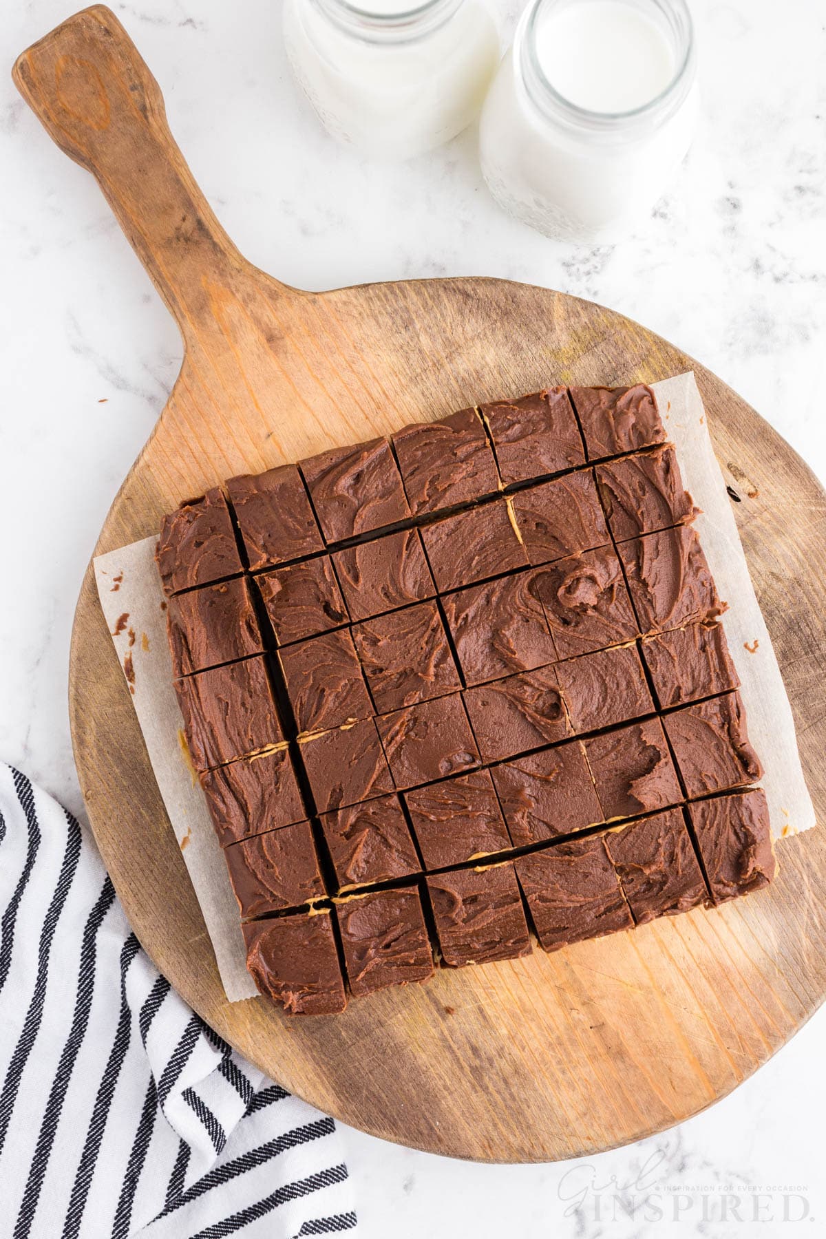 Hardened chocolate peanut butter fudge cut into square pieces on a wooden kitchen board, striped linen, two jars of milk, on a marble countertop.