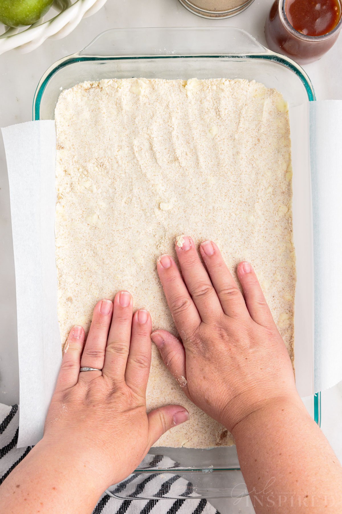 Hands pressing shortbread crust ingredients into parchment paper-lined glass baking dish.
