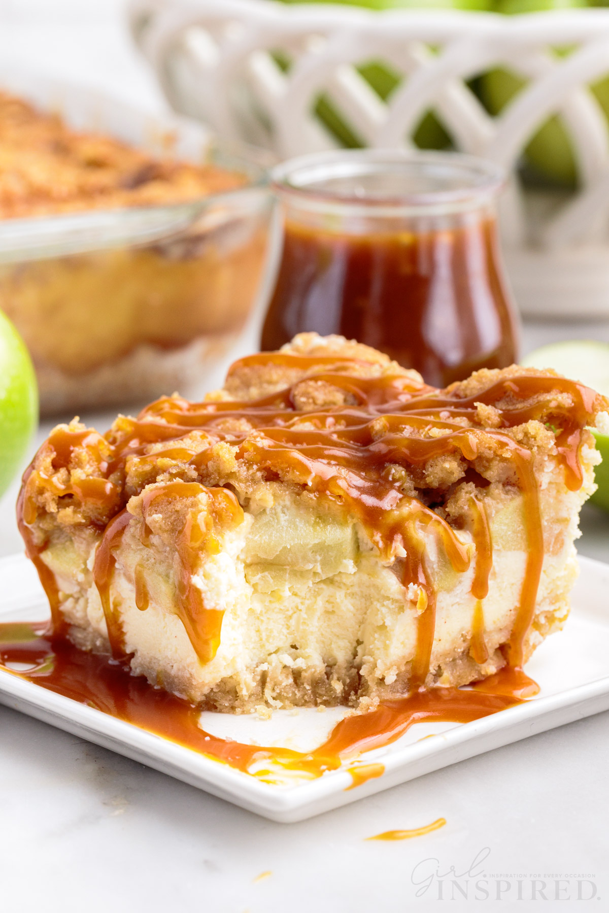 A square of apple cheesecake bar served with caramel sauce topping on a small plate with a bite removed, jar of caramel sauce.