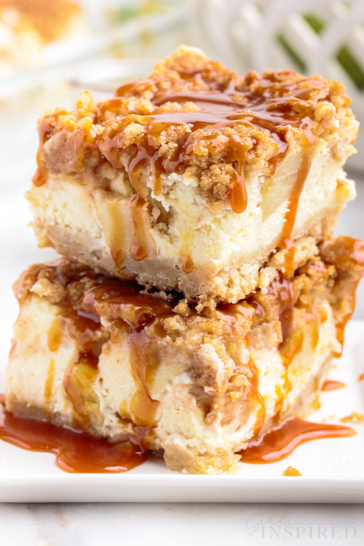Stack of two apple crumble cheesecake bars on a white plate with caramel sauce drizzled over the top.