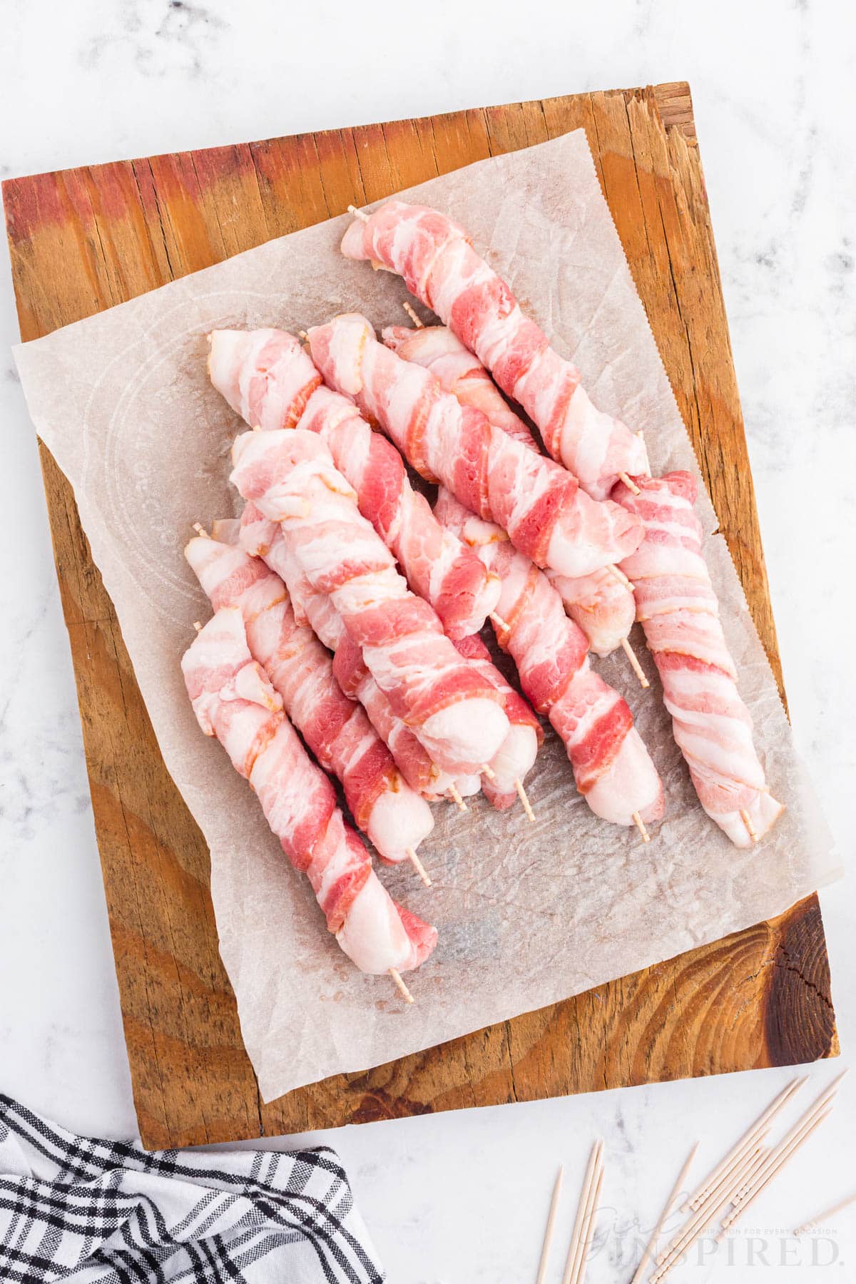 Bacon wrapped Mozzarella cheese sticks on parchment paper on a wooden kitchen board, navy checkered linen, toothpicks on a marble countertop.