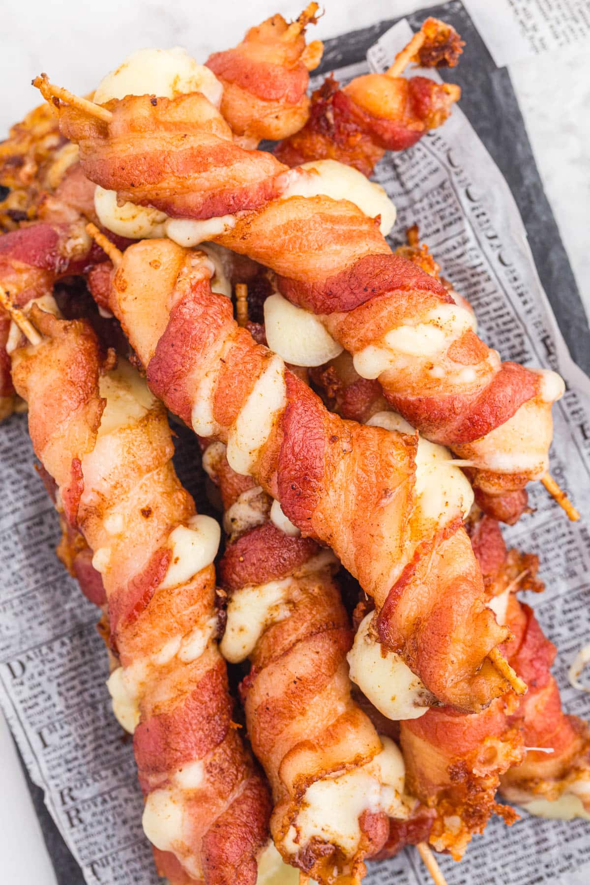 Close up of bacon wrapped mozzarella sticks on parchment paper on a black slate plate, on a marble countertop.