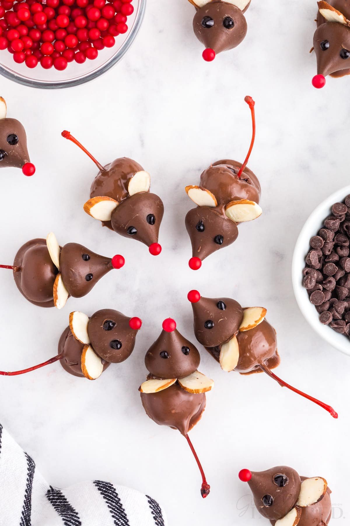 Fully assembled chocolate cherry mice on a white marble countertop, striped navy linen, bowl of red candies, bowl of chocolate chips.