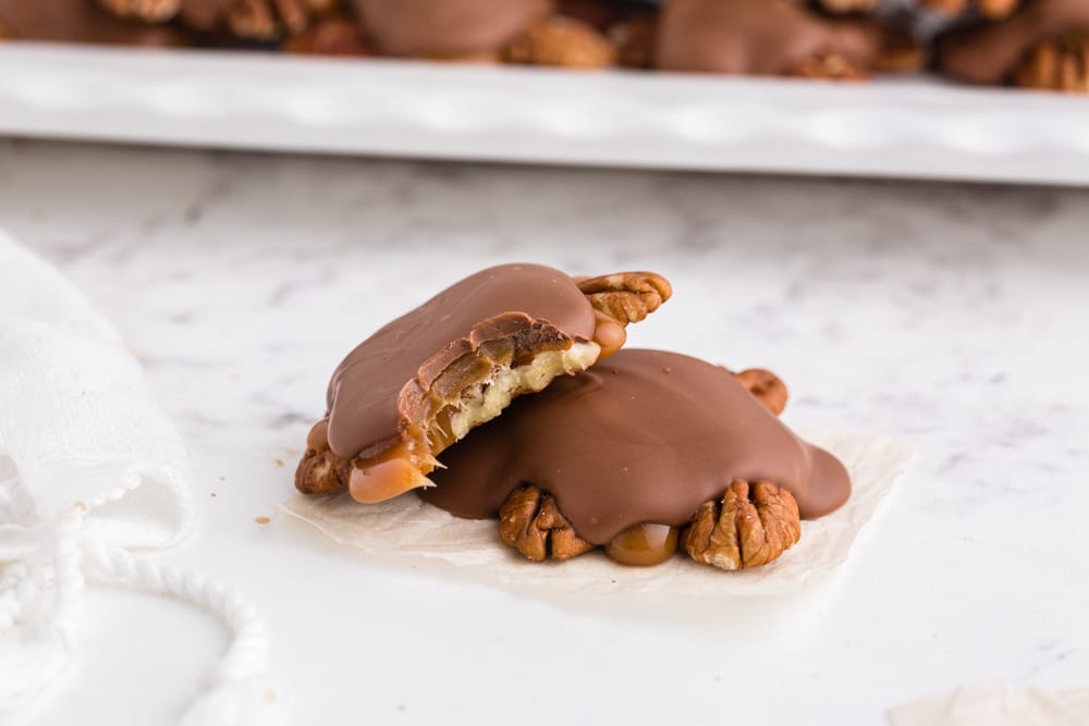Two assembled chocolate turtles, a bite taken from one chocolate turtle, tray of chocolate turtles on a white platter, on a marble countertop.