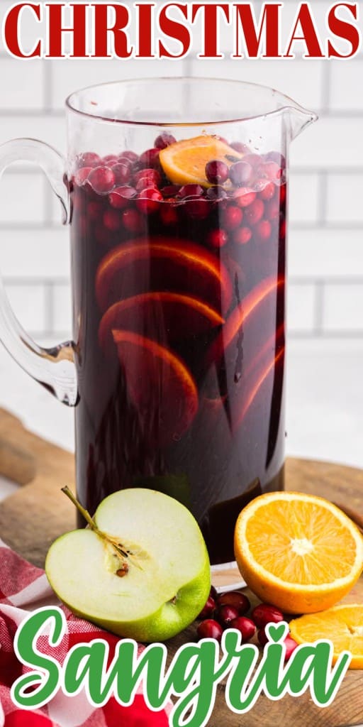 Glass Pitcher full of Red Sangria with oranges and Cranberries on a wooden kitchen board, half an apple, half and orange, red and white checked linen, white subway tiles in the background.
