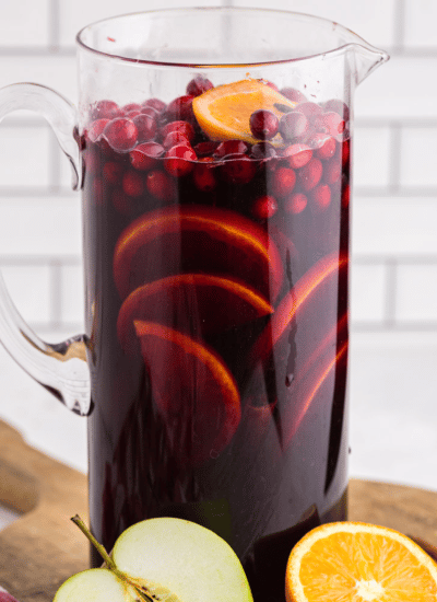 Glass Pitcher full of Red Sangria with oranges and Cranberries on a wooden kitchen board, half an apple, half and orange
