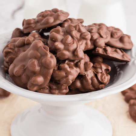 Batch of Crockpot Chocolate Peanut Butter Clusters on a cake stand, two jars of milk in the background.