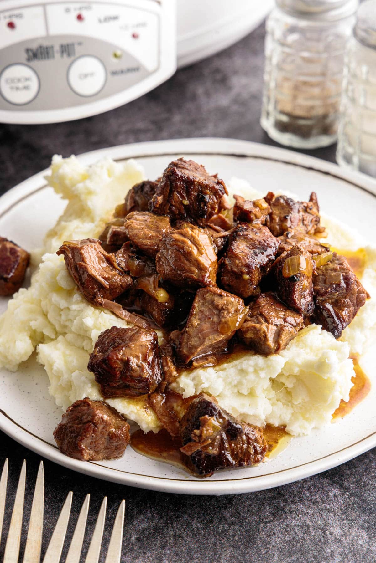crockpot steak bites on top of mashed potatoes on a plate next to forks and salt and pepper