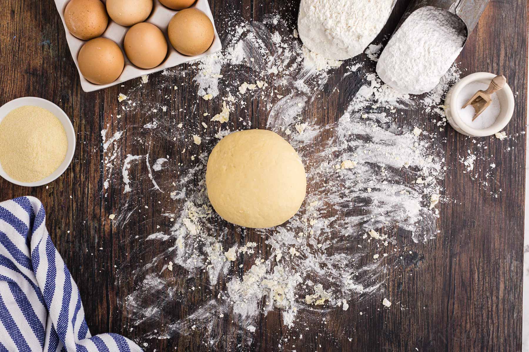 Homemade pasta dough rolled into a smooth ball on a wooden kitchen surface, bowl of semolina, striped linen, tray of eggs, small bowl of slat, measuring cups with different types of flour.