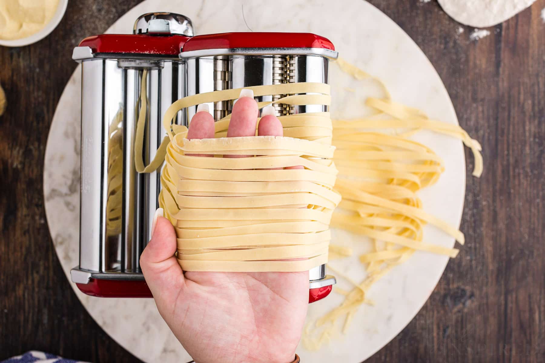 Hand holding thin fettucine pasta strips over a pasta machine on a marble board, bowl of semolina, flour, on a wooden kitchen countertop.