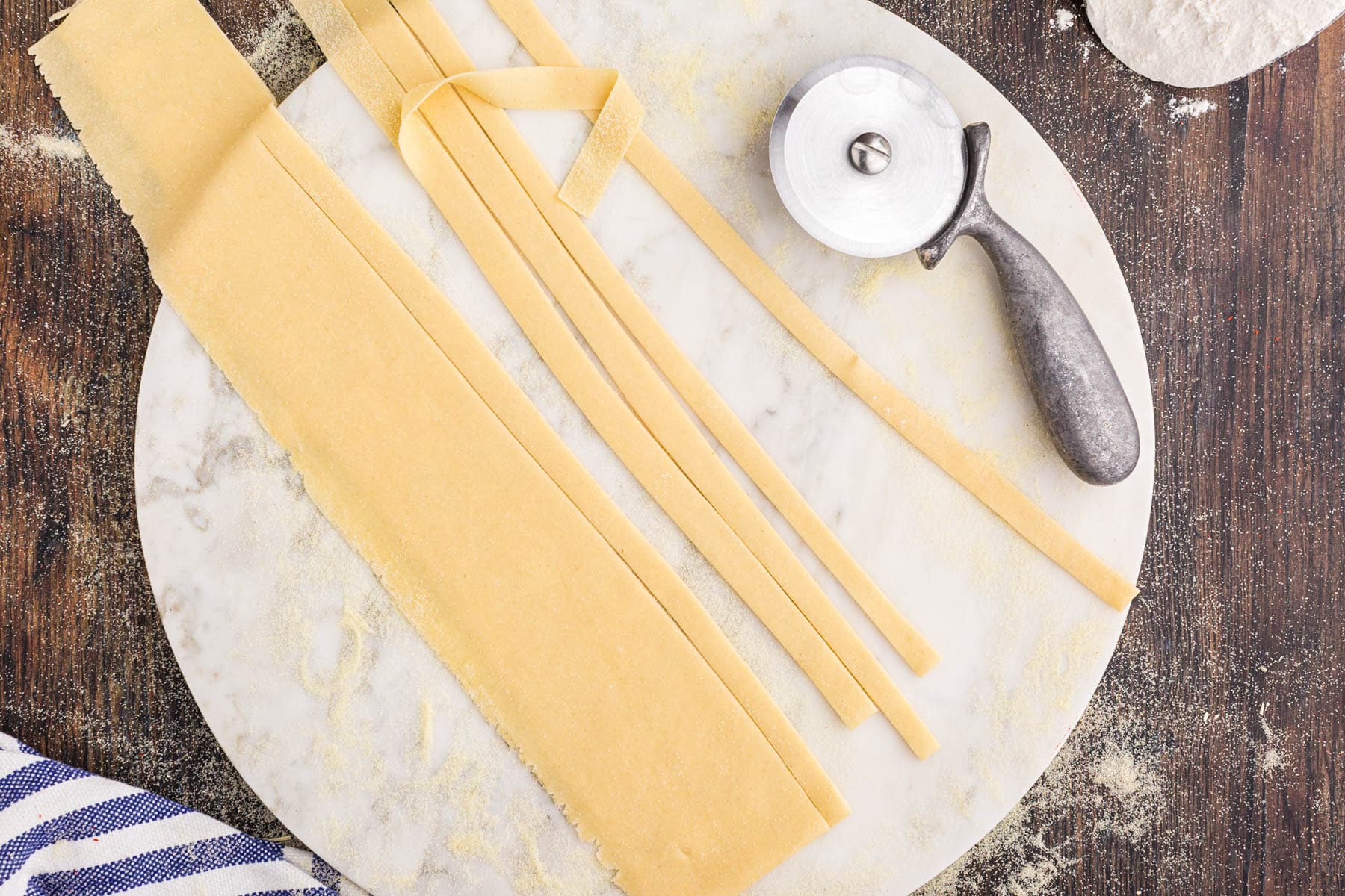 Thin fettuccine strips of pasta dough on a marble board with metal pizza cutter, striped linen, flour, on a wooden kitchen countertop.