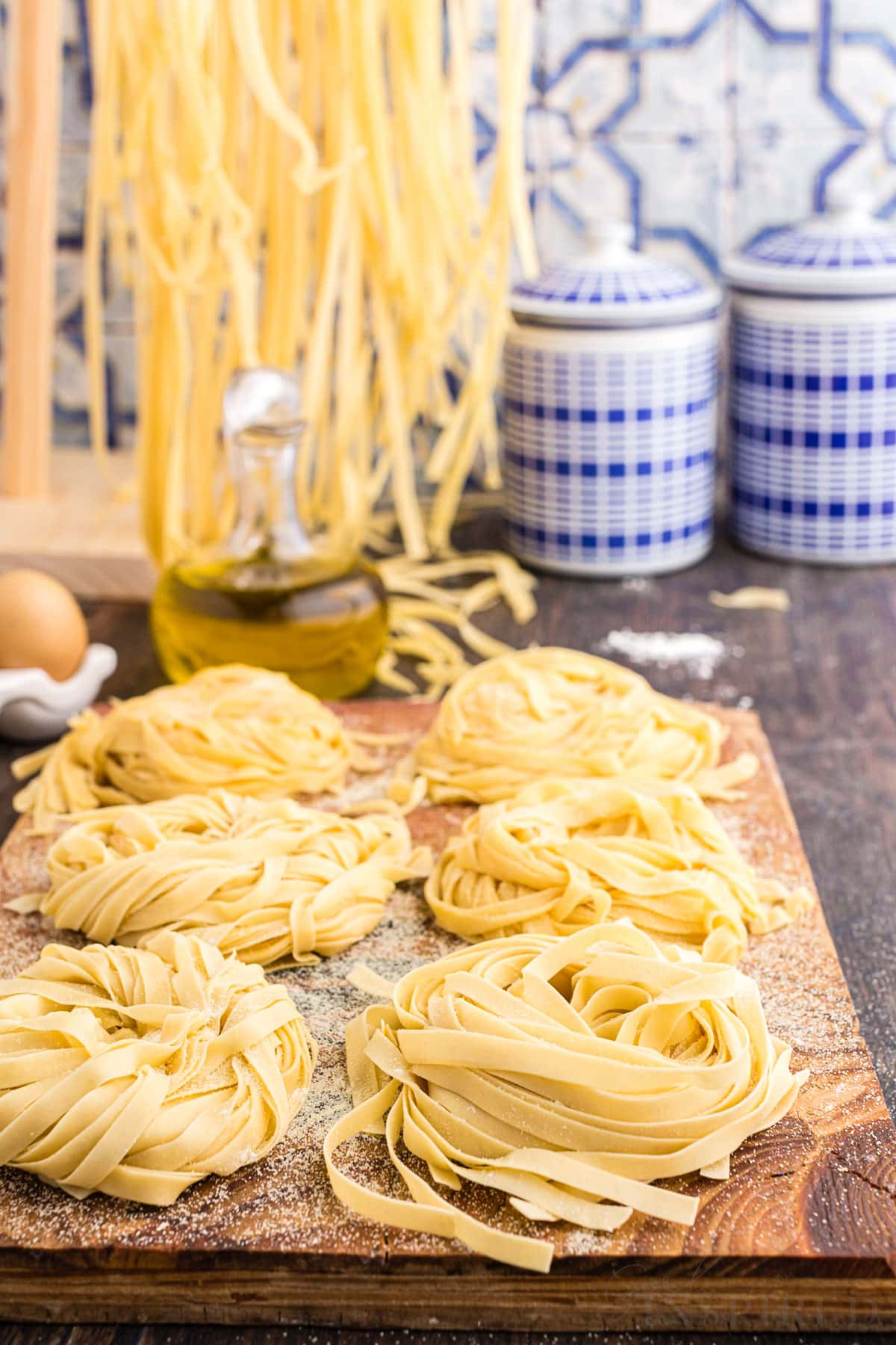Homemade pasta on a wooden kitchen board, pasta strips hanging from a pasta machine, blue decorative ingredients jars in the background with blue patterned tiled wall.