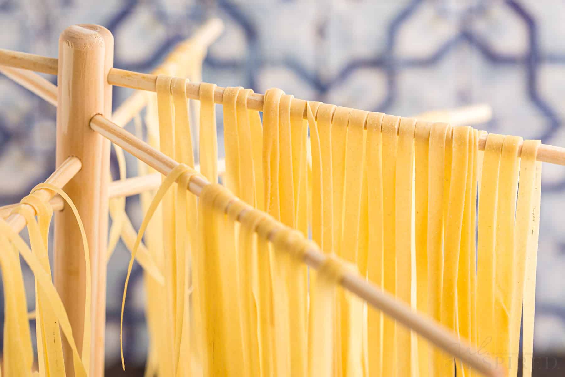 Thin fettuccine strips of homemade pasta hanging to dry on a pasta rack, blue and white tiles in the background.