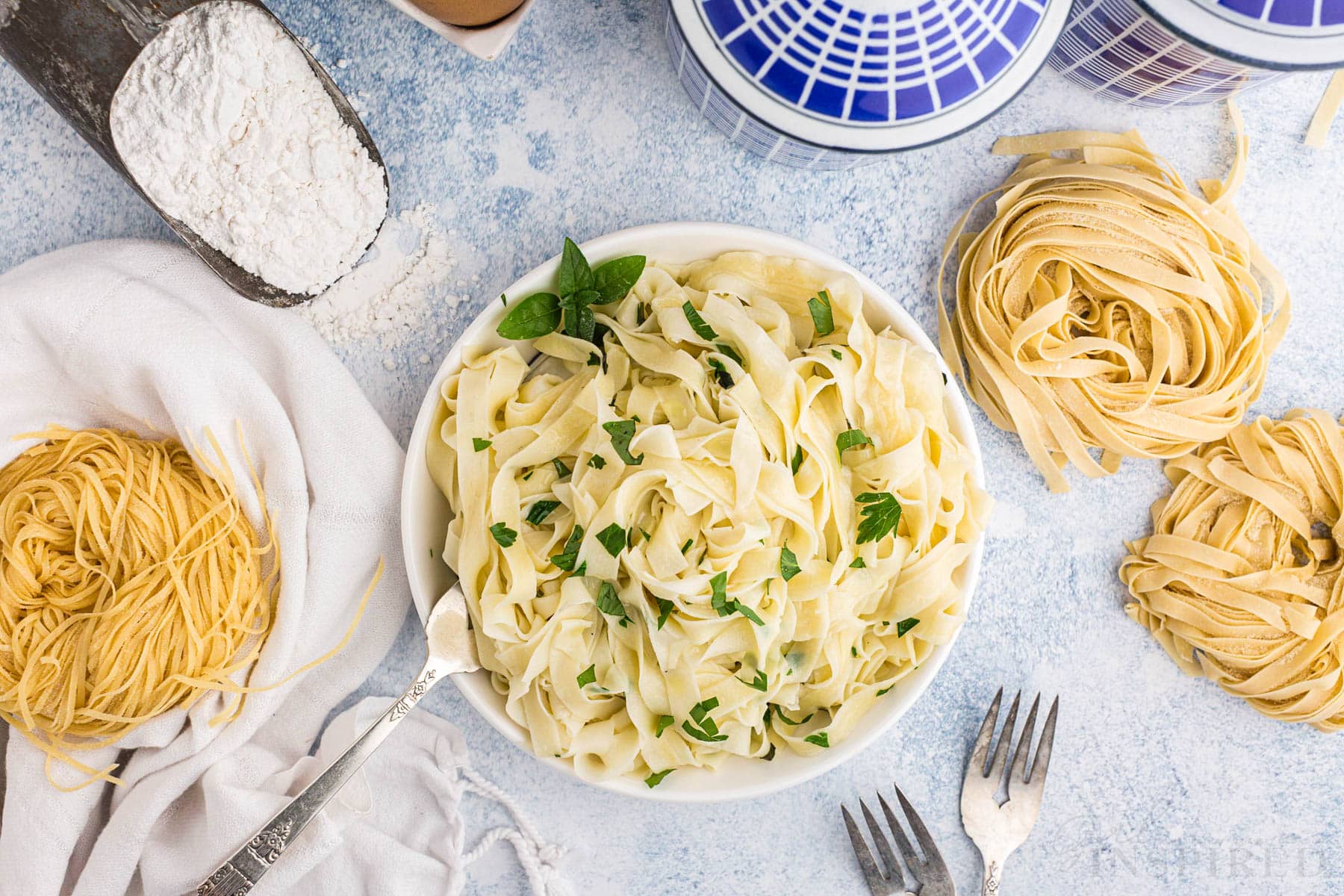 Overhead of bowl of cooked homemade pasta with fresh herbs and metal fork, two metal forks on the countertop, metal measuring spoon with flour, cluster of uncooked homemade pasta, white linen, blue decorative ingredient jars, on a marble countertop.